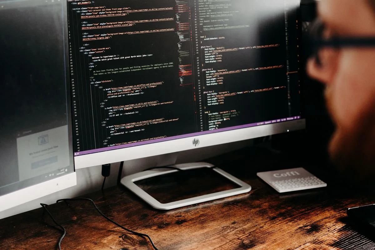 Person mit Brille vor einem Monitor mit farbig hervorgehobenem Quelltext, daneben eine Tastatur auf einem Holzschreibtisch