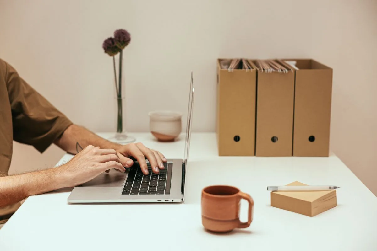 Person arbeitet am Laptop an einem hellen Schreibtisch mit Kaffeetasse
